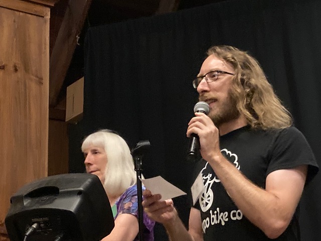 A picture of Sam standing in front of a monitor holding a Mic and card at the John C. Campbell Folk School while Gaye Phifer looks on in the background.