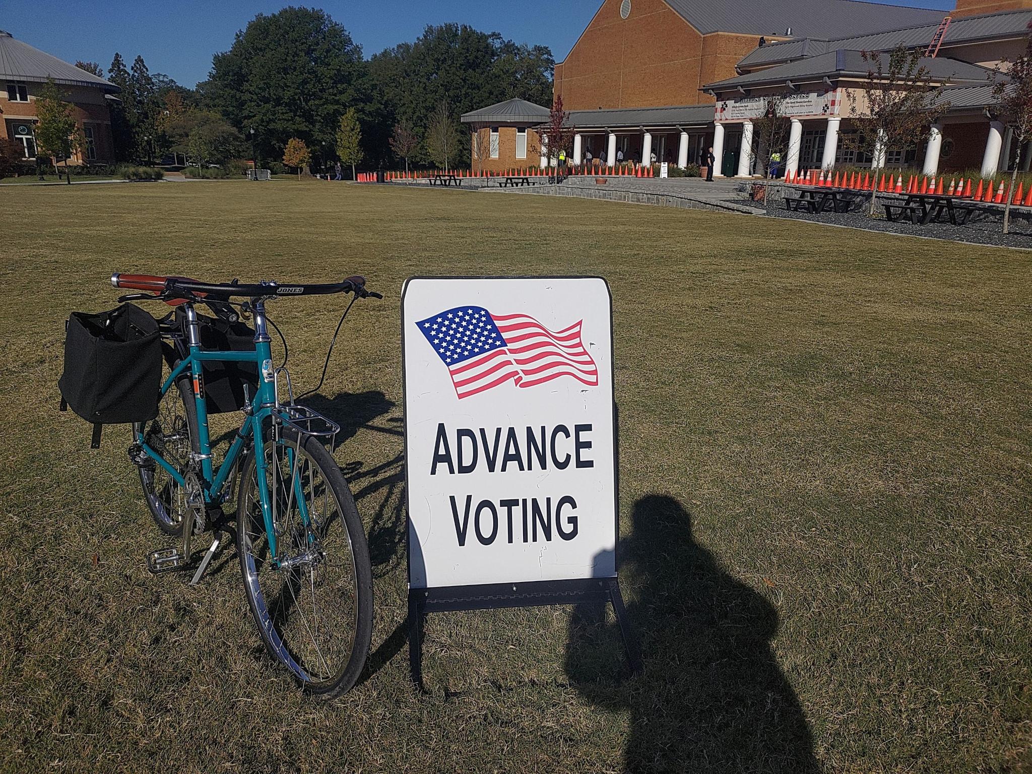 A blue bicycle next to a sign that says 'Advance Voting' and has a picture of a U.S. flag. Behind it is a brick building with a short line out front and lots of cones set out to direct the line away from doors.