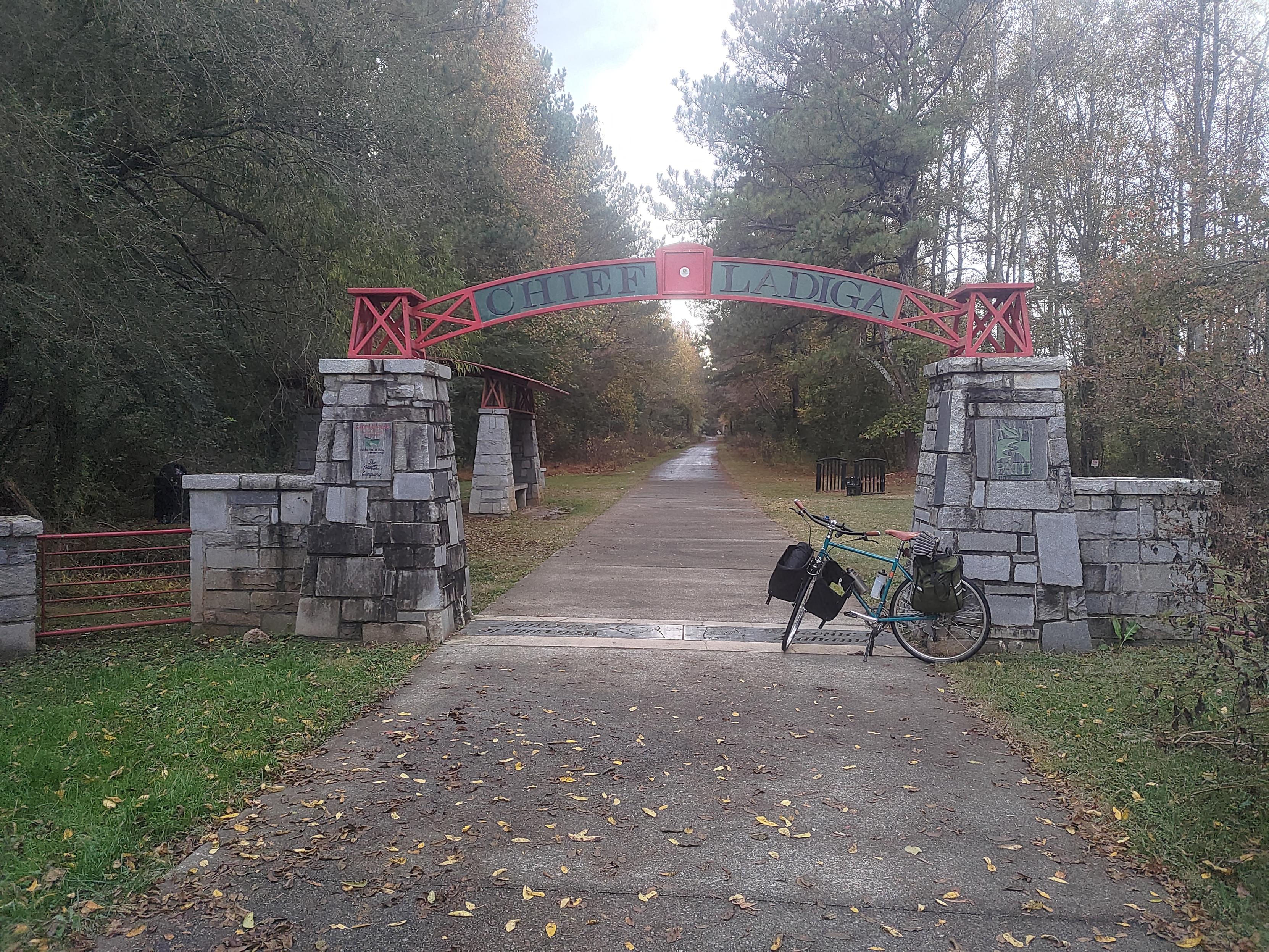 Two stacked stone pillers with a big red metal arch on to that says 'Chief Ladiga'. The bike path runs through the arch and a bicycle sits under it.