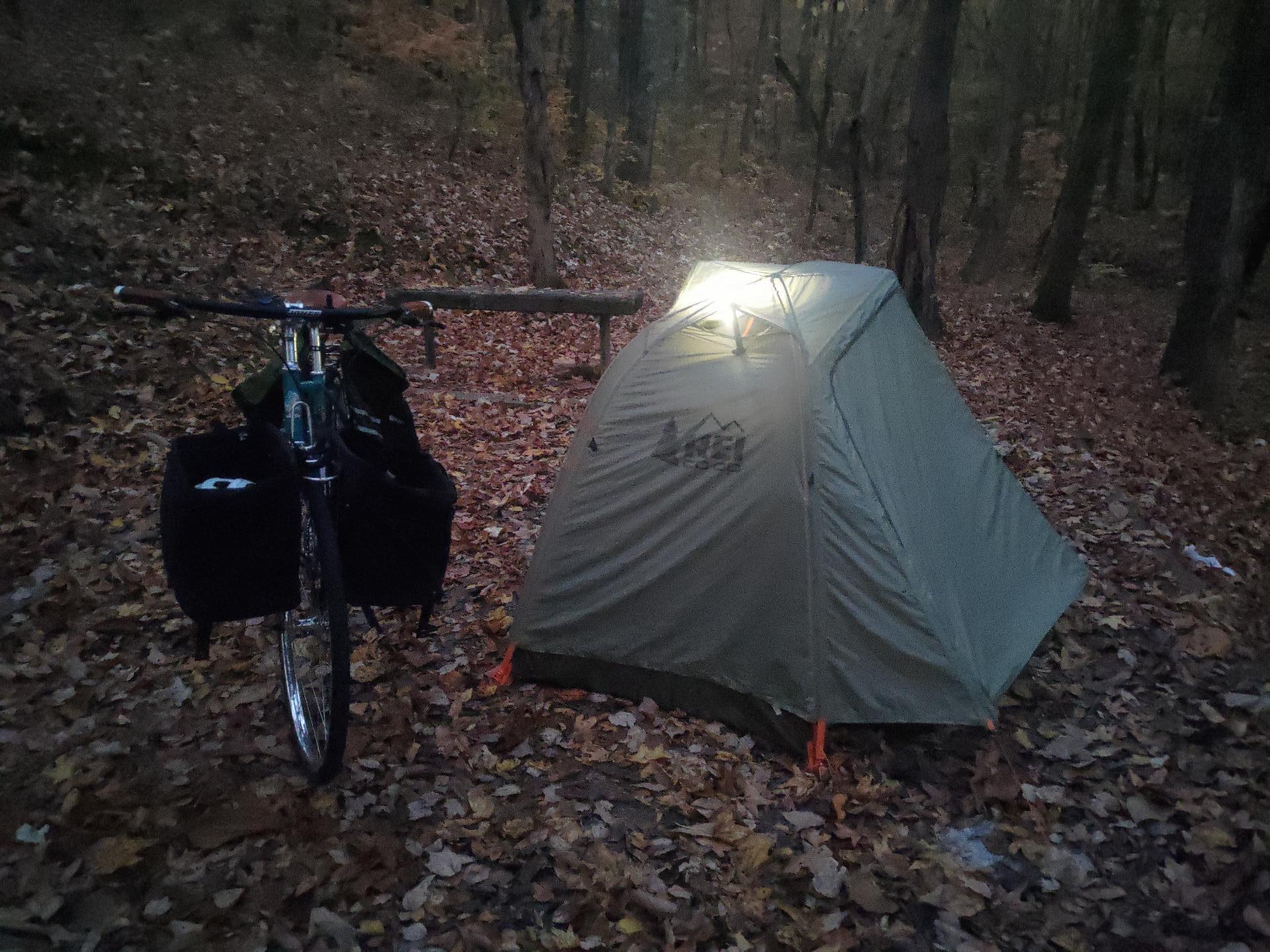 A small green tent with a light on inside that shines through the rain fly next to a blue bicycle with panniers. It is dusk and the light is low and it's all a bit hard to see.