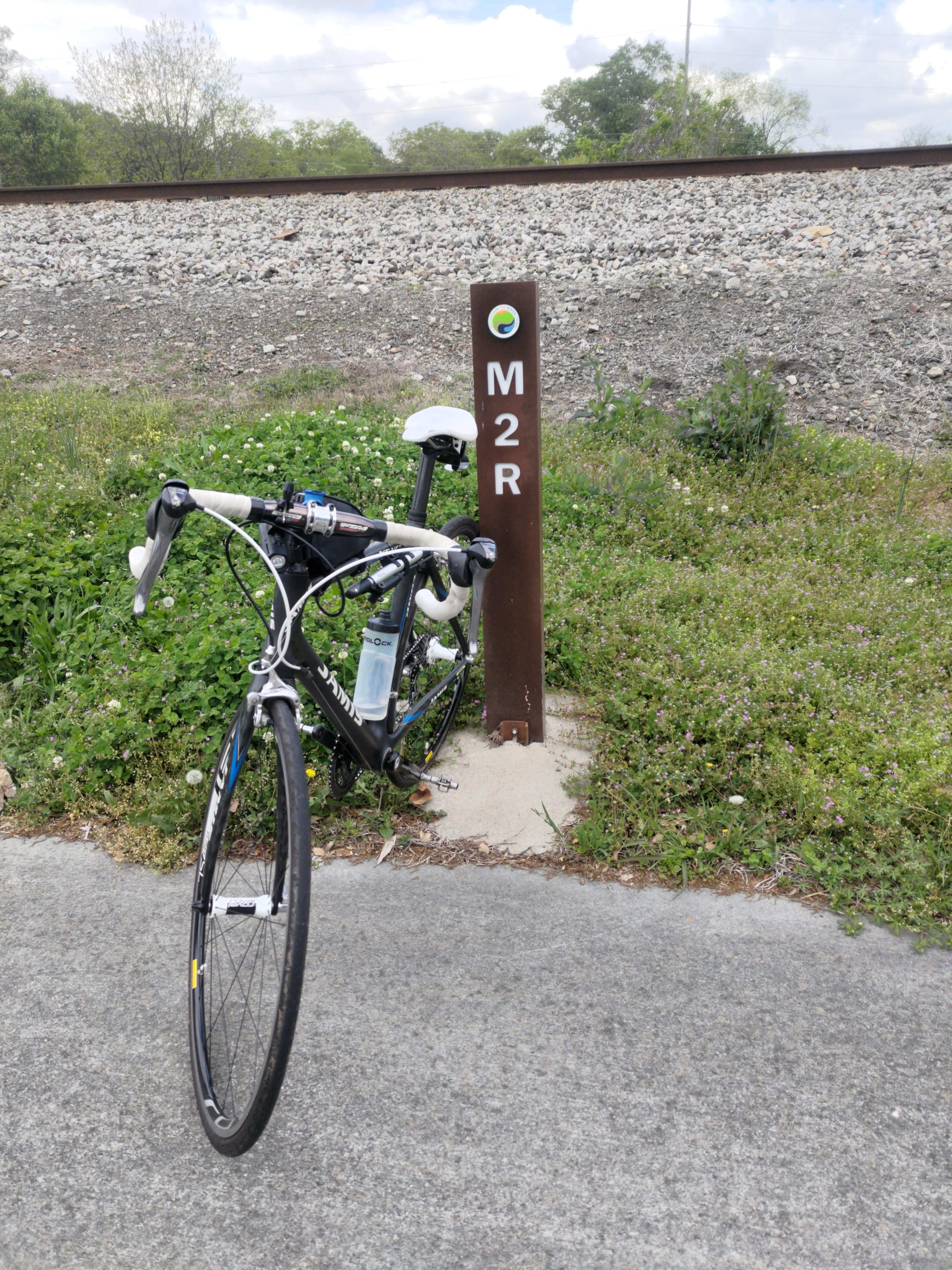 A black and white road bike sits propped against a trail marker that says 'M2R', there are some train tracks up an embankment in the background